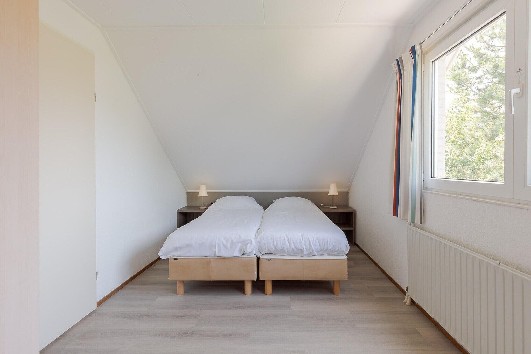 Une chambre avec un plafond en pente, deux lits simples, des tables de chevet avec des lampes, une fenêtre avec des rideaux et un radiateur.