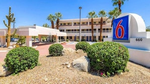 Un motel con un letrero azul llamativo que muestra el número 6, un edificio de varios pisos con techo plano, y un estacionamiento con palmeras y arbustos.