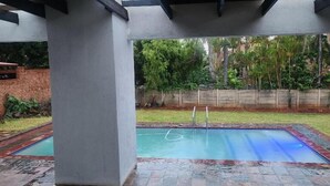 A covered outdoor swimming pool with a tiled deck, a metal ladder, and a brick wall in the background. - Cozy and Modern Apartment Rental in Akasia, Pretoria (Akasia)