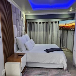 Un dormitorio moderno con una cama grande, una mesita de noche, un televisor de pantalla plana y vistas a un atardecer a través de cortinas.