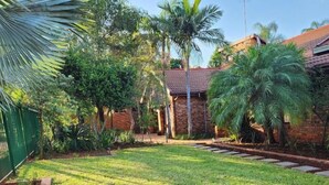 A well-maintained garden with a lawn, palm trees, and a brick building in the background. - Cozy and Modern Apartment Rental in Akasia, Pretoria (Akasia)
