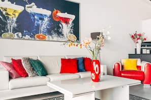A modern living room with a white sofa, red and blue cushions, a red vase with numbers, and a black and white abstract painting above.