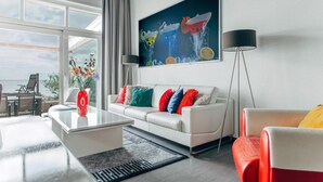 A modern living room with a white sofa, colorful cushions, a glass coffee table, a red vase, and a large painting on the wall. - Deluxe Dike Villa with Sauna for 6 (Makkum)