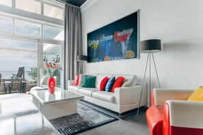 A modern living room with a white sofa, colorful cushions, a glass coffee table, a red vase, and a large painting on the wall. - Deluxe Dike Villa with Sauna for 6 (Makkum)