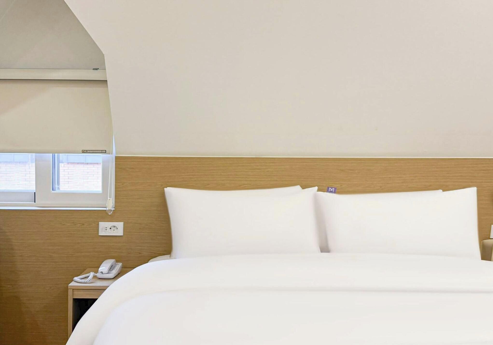 A hotel room with a bed, a bedside telephone, and a window.