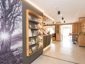 Eine moderne Hotelhalle mit einem Holzbüro, einem Bücherregal mit Zeitschriften und einer großen Wandmalerei mit Baumotiv.