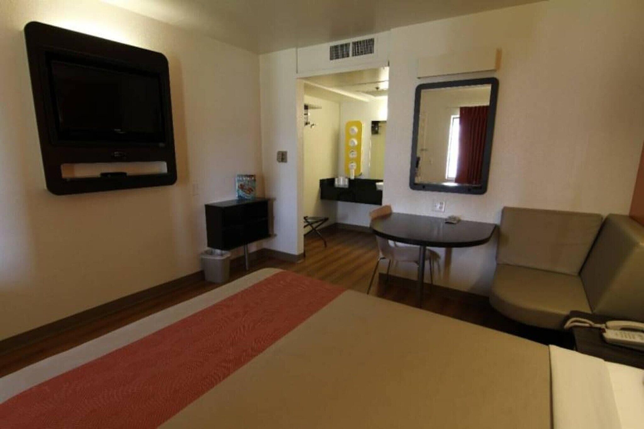 A hotel room with a bed, a TV mounted on the wall, a small table with chairs, a sofa, and a mirror.