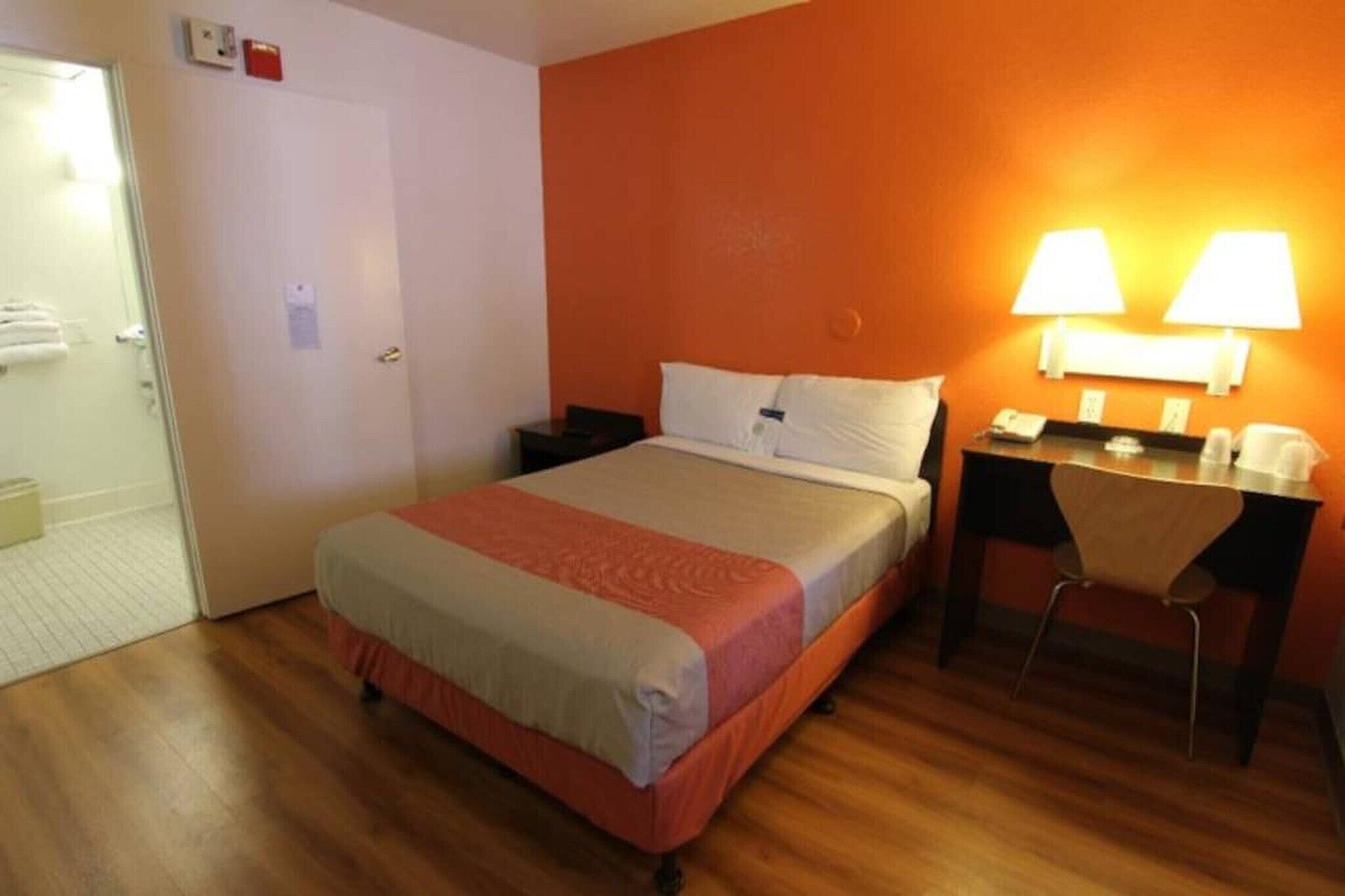 A hotel room with a bed, bedside table, desk, chair, and a door.