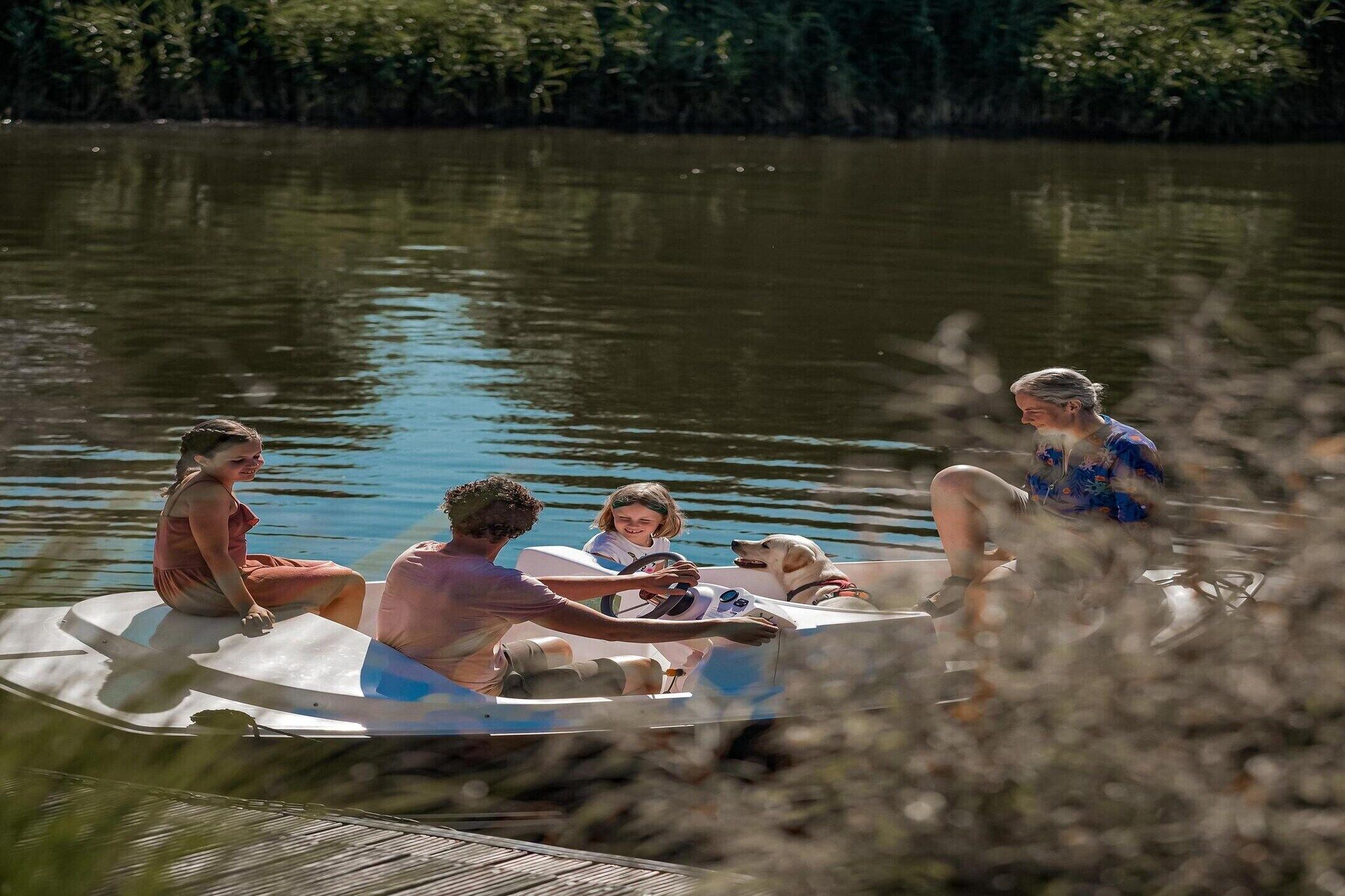 A group of people on a small boat with a dog, surrounded by calm water and greenery.