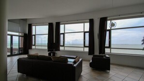 Una sala de estar con un sofá, una mesita y un televisor. Hay ventanas grandes con cortinas, y vistas al mar.