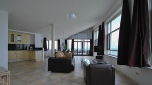 A spacious living area with a sofa, a coffee table, and a television. There is a kitchen area with cabinets and a dining table. Large windows provide a view of the outdoors. - Lakeview Apartment with Dual Balconies (Makkum)