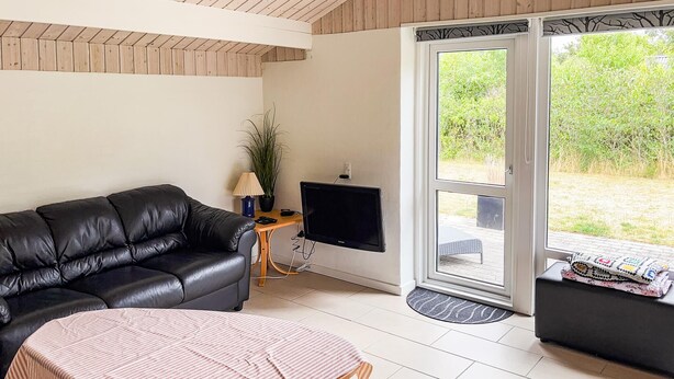 A living room with a black leather sofa, a small wooden table with a lamp, a television mounted on the wall, and a view of a garden through a glass door. - 3 bedroom nice home in Skjern (Skjern)