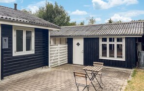 A building with a dark exterior, a white door, and a window. There is a small table and a bench in the foreground. - 3 bedroom nice home in Skjern (Skjern)
