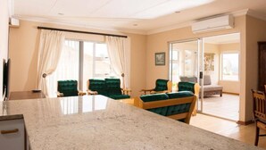 Un vaste espace de vie comprenant un grand îlot de cuisine, des chaises recouvertes d’un tissu vert et une porte-fenêtre coulissante donnant sur un balcon.