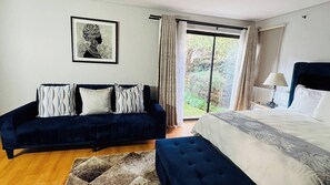 Ein Schlafzimmer mit einem Bett, einem dunkelblauen Sofa, einem gerahmten Kunstwerk und Blick durch ein Fenster.