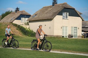 Dos personas andando en bicicleta por un sendero, con edificios de techo de paja al fondo.