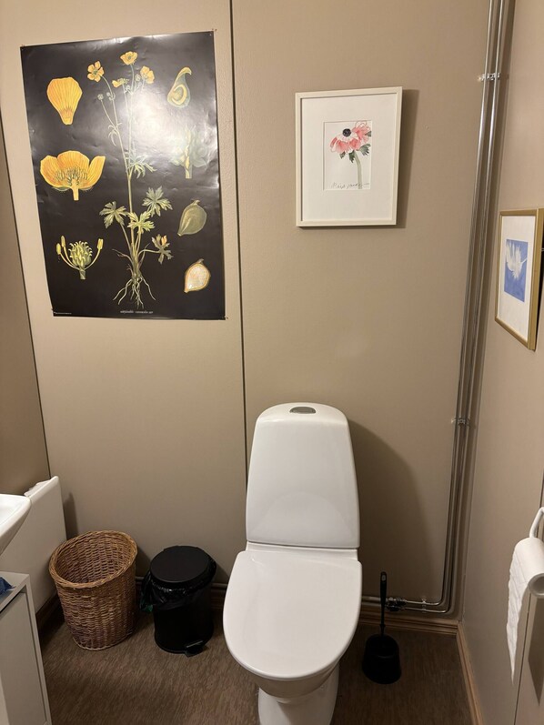 Un baño pequeño con inodoro, un cubo de basura, una cesta y una ilustración botánica enmarcada en la pared.