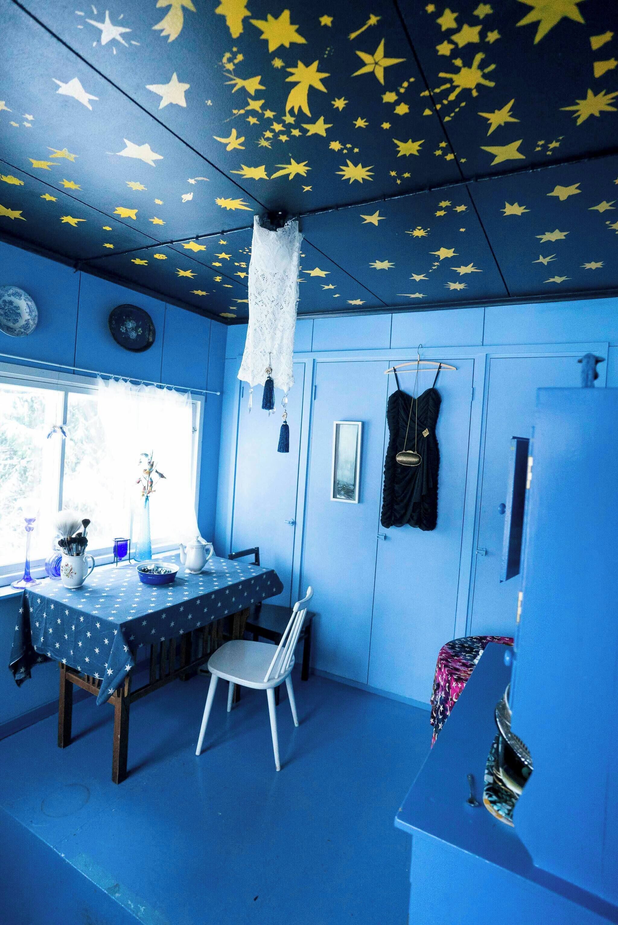 A room with a star-patterned ceiling, a table with a polka dot tablecloth, a white chair, and a black dress hanging on the wall.