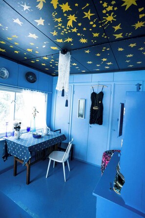 Ein Zimmer mit einer sternenmusterigen Decke, einem Tisch mit einem punktierten Tischläufer, einem weißen Stuhl und einem schwarzen Kleid, das an der Wand hängt.