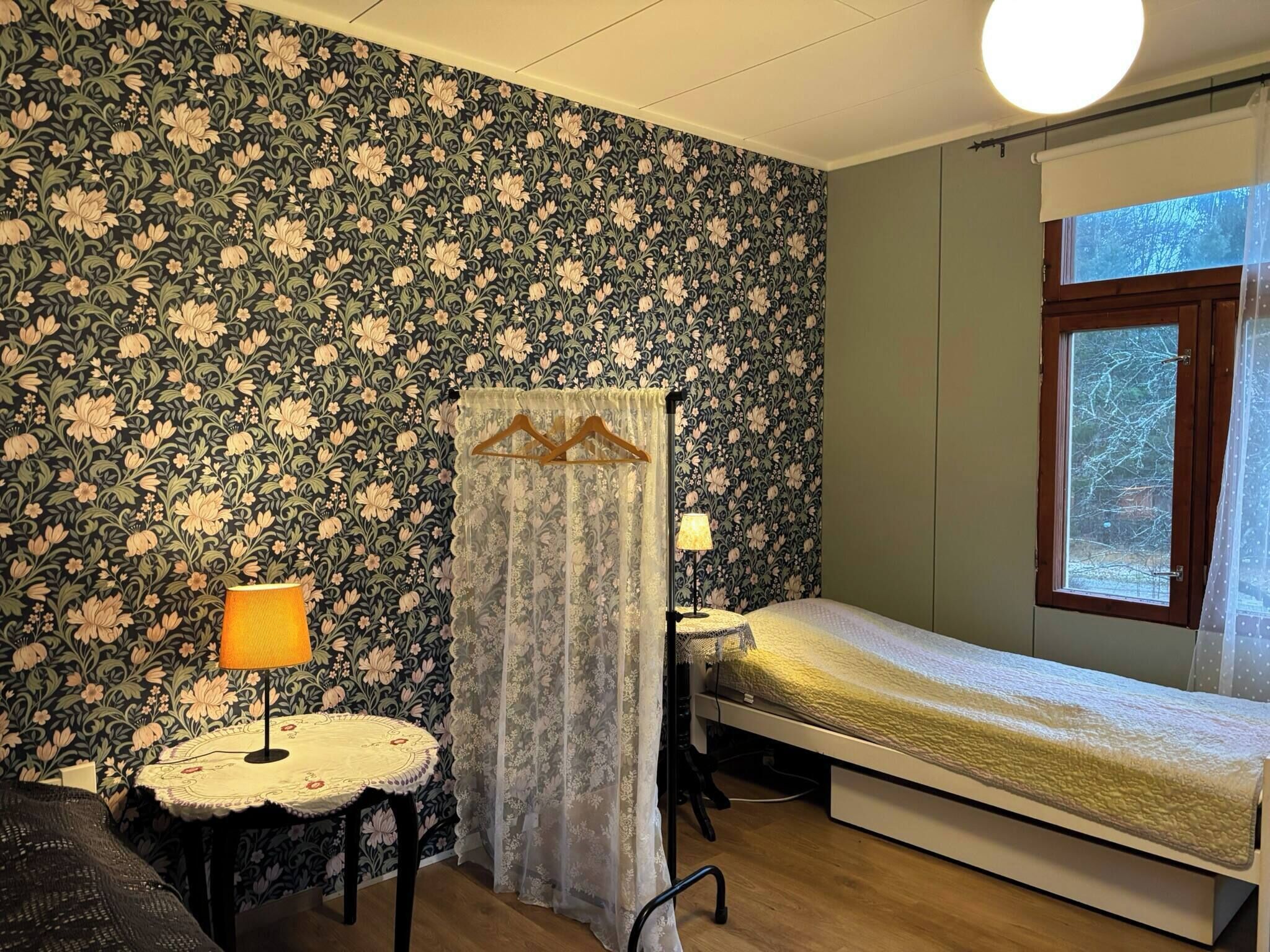 A room with floral wallpaper, a bed, a small table with a lamp, and a window with a view of trees.