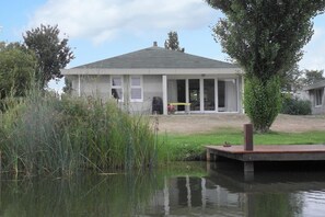 A house with a covered porch, a pond in the foreground, and a grassy area with a tree. - Elegant Waterfront Bungalow with Jetty (Makkum)