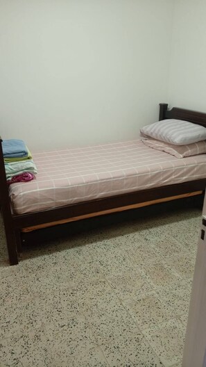 Ein Einzelbett mit Holzrahmen, einer Matratze und einem Kopfkissen in einem Zimmer mit schlichten Wänden und punktierten Bodenfliesen.