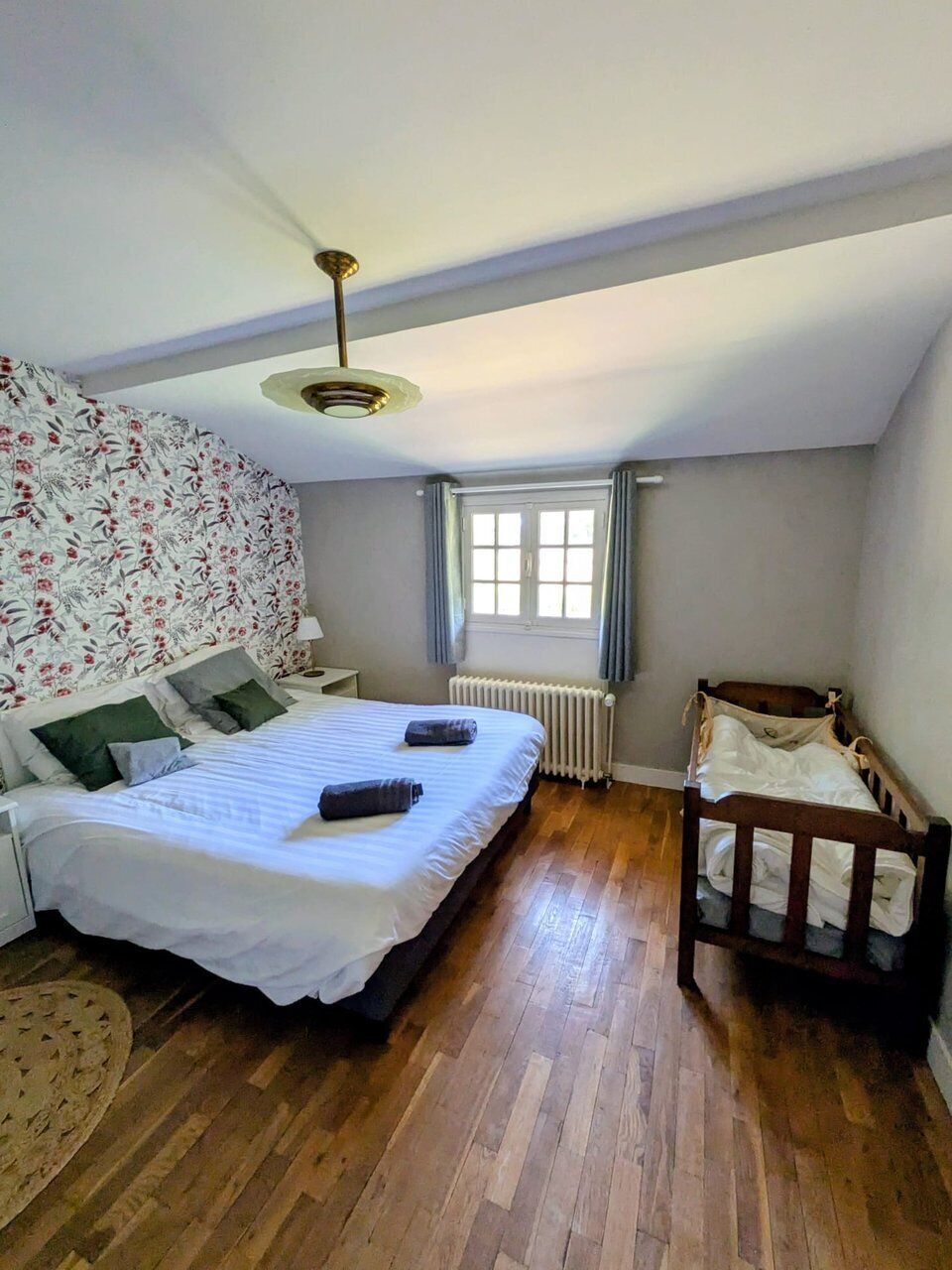 Une chambre à coucher avec un grand lit, un lit plus petit, un berceau en bois, un radiateur, une fenêtre avec des rideaux et un ventilateur de plafond.