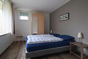 A bedroom with a bed, a chair, a bedside table, a lamp, a window with curtains, a door, and a wall-mounted picture. - Comfortable Bungalow Stay by the Water (Makkum)