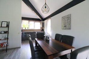 A modern dining area with a long table, black chairs, a hanging light fixture, and a kitchen in the background. - Comfortable Bungalow Stay by the Water (Makkum)