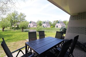 A covered outdoor seating area with a table and chairs, overlooking a pond and houses in the background. - Comfortable Bungalow Stay by the Water (Makkum)