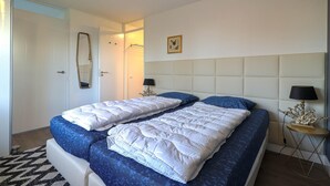 Ein Doppelbett mit weißer und blauer Bettwäsche, ein Nachttisch mit Lampe sowie ein an der Wand befestigter Spiegel.
