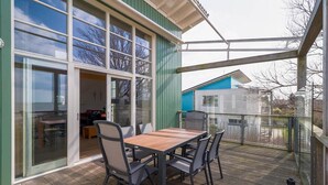 Eine überdachte Terrasse mit Holztisch und -stühlen, großen Schiebeglastüren und Blick auf ein blaues Gebäude im Hintergrund.