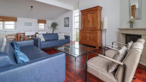 Ein geräumiges Wohnzimmer mit einem blauen Sofa, einem beigen Sessel und einem Glastisch. Im Hintergrund befindet sich ein Holzschrank und ein Kamin.