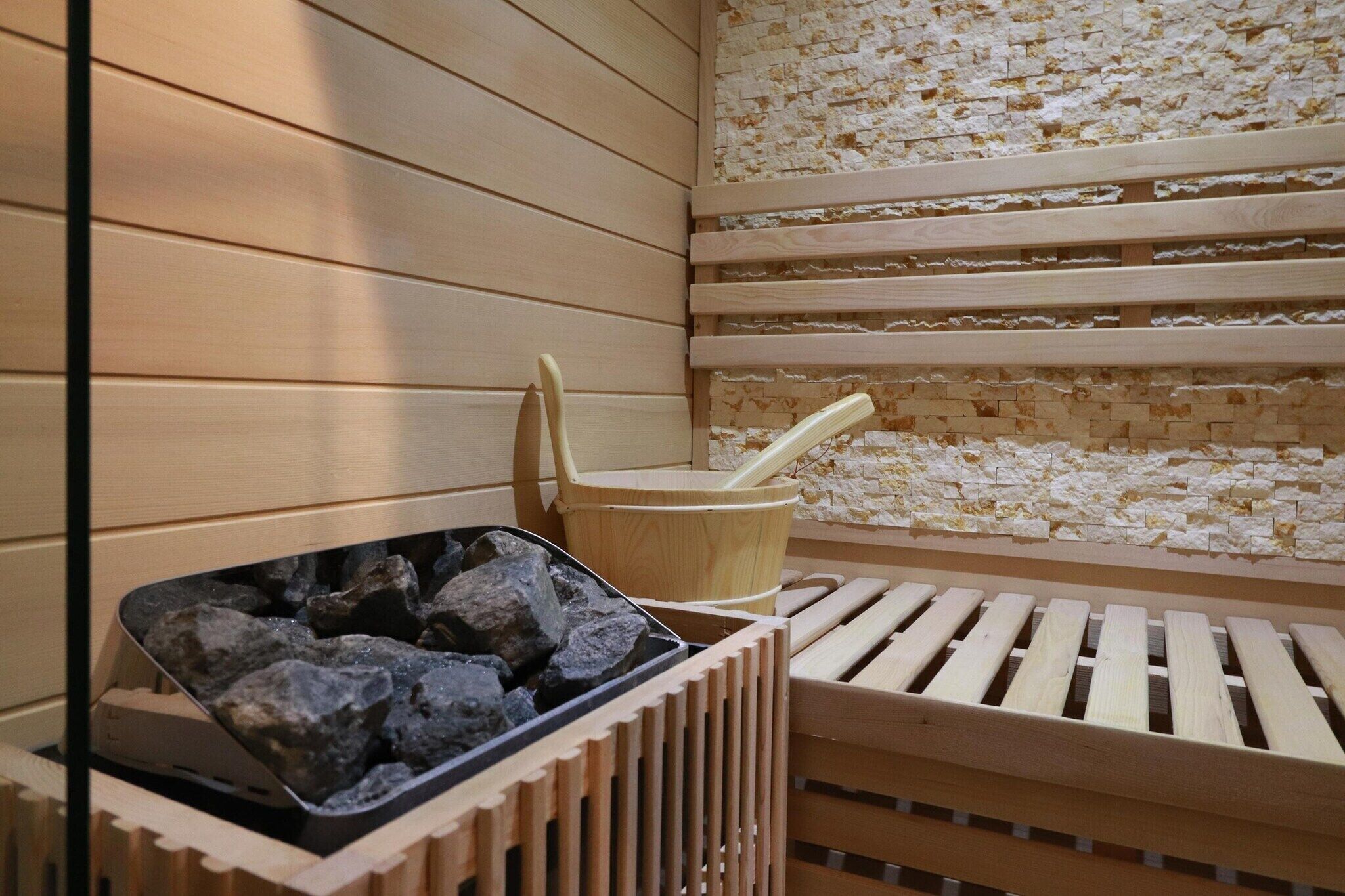 Eine Sauna mit Holzbänken, einer Steinwand und einem Eimer mit Wasserschöpflöffel.