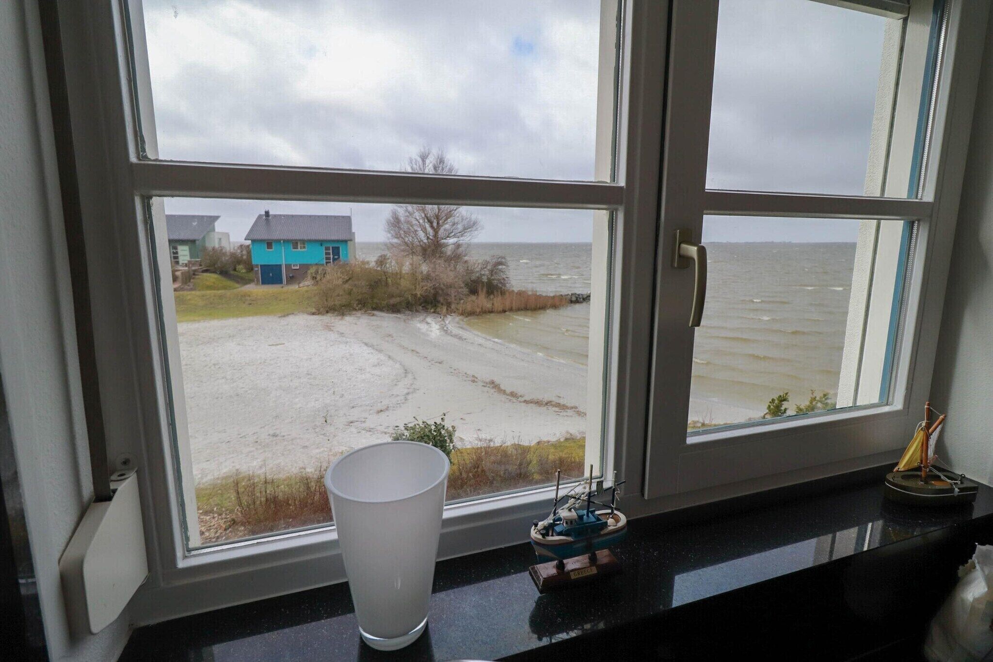 Une vue depuis une fenêtre donnant sur une plage de sable, avec une petite maison et un modèle de bateau sur le rebord de la fenêtre.