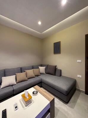 A modern living room with a grey sectional sofa, a small wooden coffee table, a remote control, and a tissue box. - 302 AL-NAJEH JRE Apartments For Rent ave (Amman)