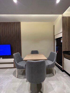 A modern kitchen with a wooden dining table, two grey upholstered chairs, a built-in TV, and a refrigerator. - 302 AL-NAJEH JRE Apartments For Rent ave (Amman)
