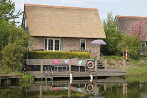 A traditional house with a thatched roof, a wooden dock, and a small boat. - Deluxe Beach Villa with Garden (Makkum)