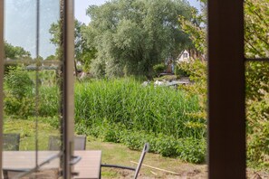 A view through a window of a garden with a pond, trees, and a building in the background. - Beach Villa with Garden & Water Access (Makkum)