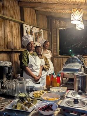 Uma cozinha com chefs usando aventais e chapéus de chef, uma mesa de buffet com diversos pratos, e uma parede de madeira com um painel decorativo.