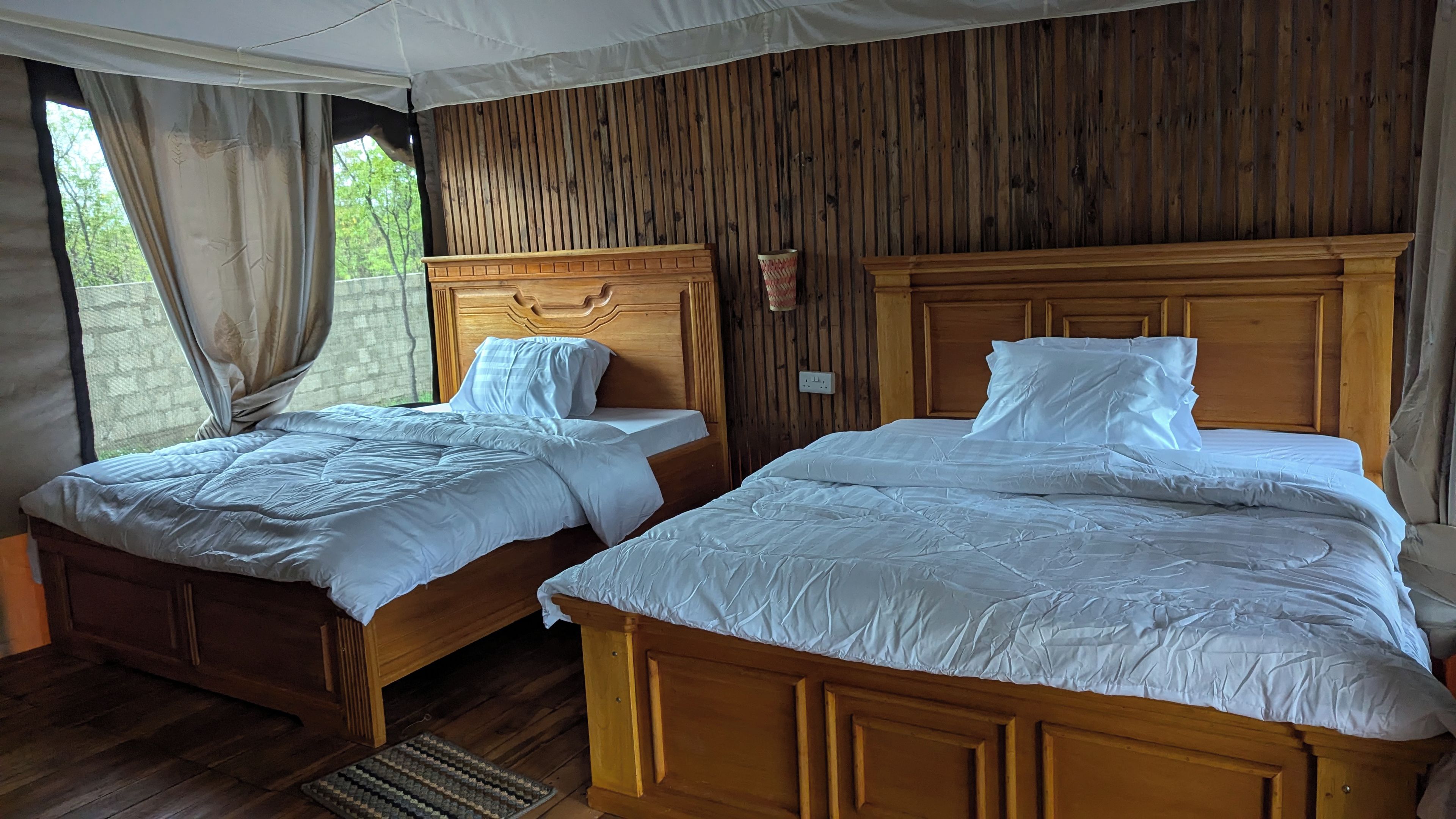 Una habitación con dos camas de madera, piso de madera, una ventana con cortinas y un panel de madera en la pared.