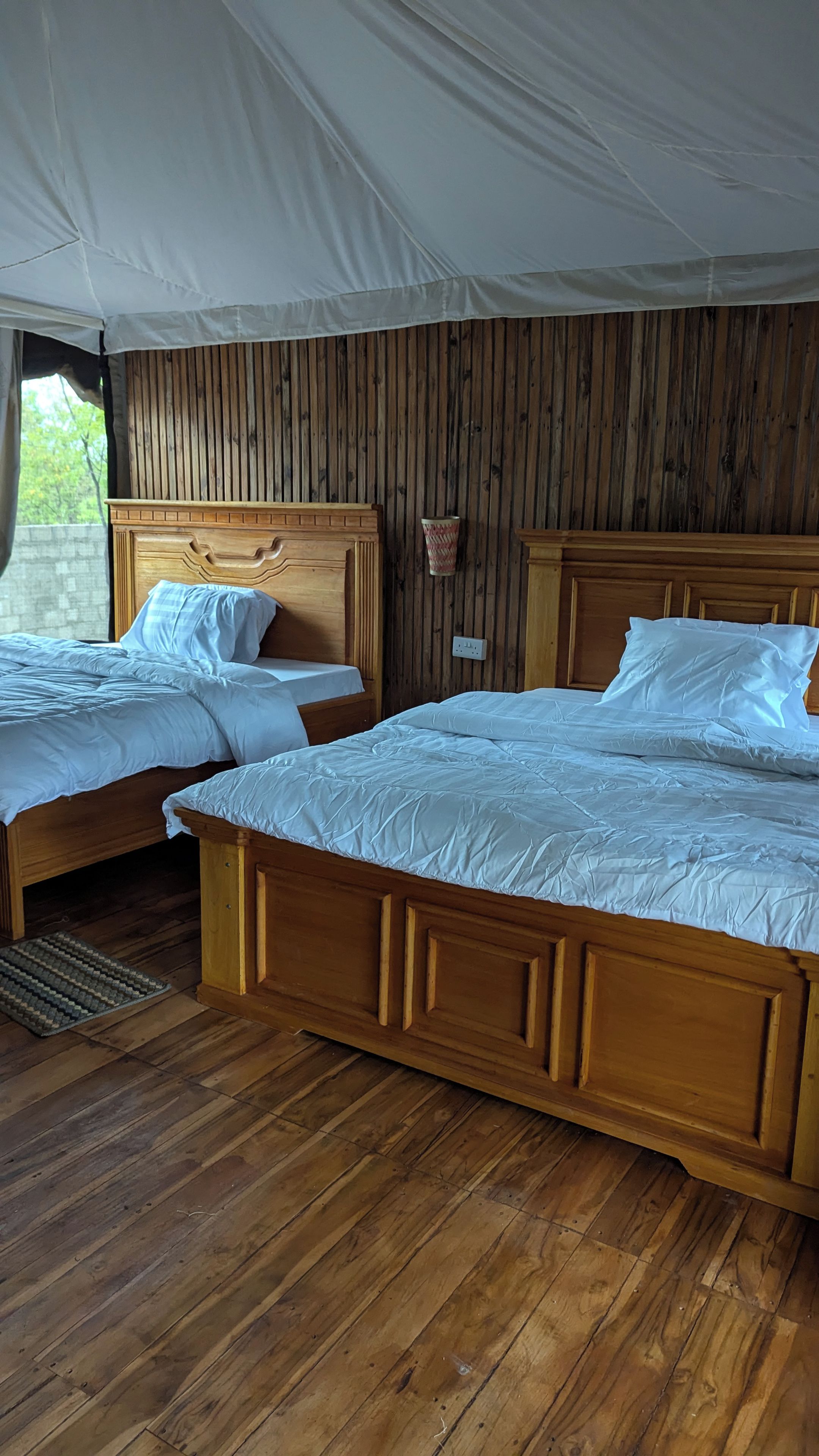 A tented accommodation with two wooden beds, a wall-mounted light, and a view of the outdoors.
