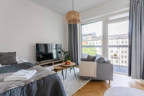 Moderni hotellihuone, jossa on sänky, sohva, televisio, pieni pöytä hedelmäkulholla sekä suuri ikkuna, josta avautuu näkymä kaupunkiin.