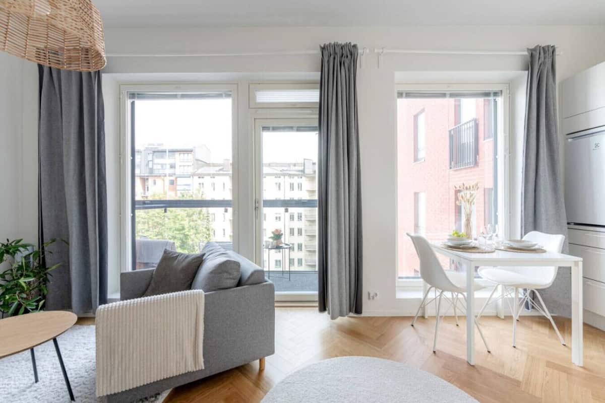 A modern apartment with a grey sofa, a dining table with chairs, and large windows with grey curtains.