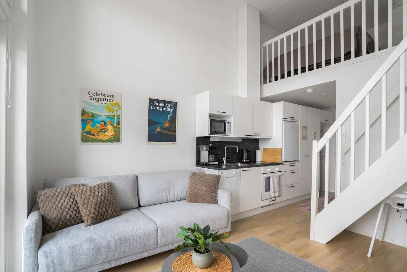 A modern living space with a grey sofa, a kitchen area, and a staircase leading to an upper level.