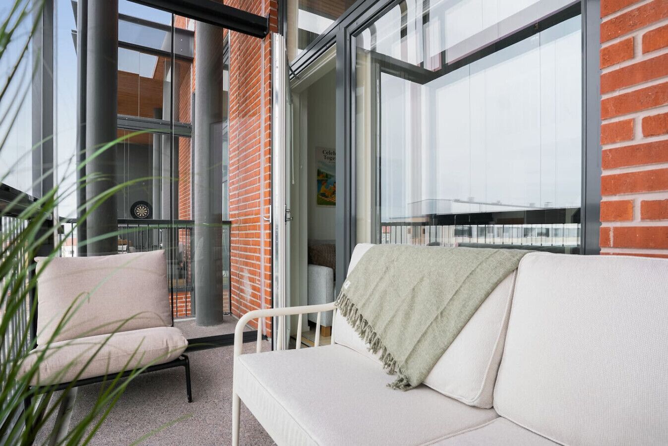 Un área de estar al aire libre y de estilo moderno con un sofá blanco y una silla, un ventanal de vidrio que da a una habitación, y un muro de ladrillo.