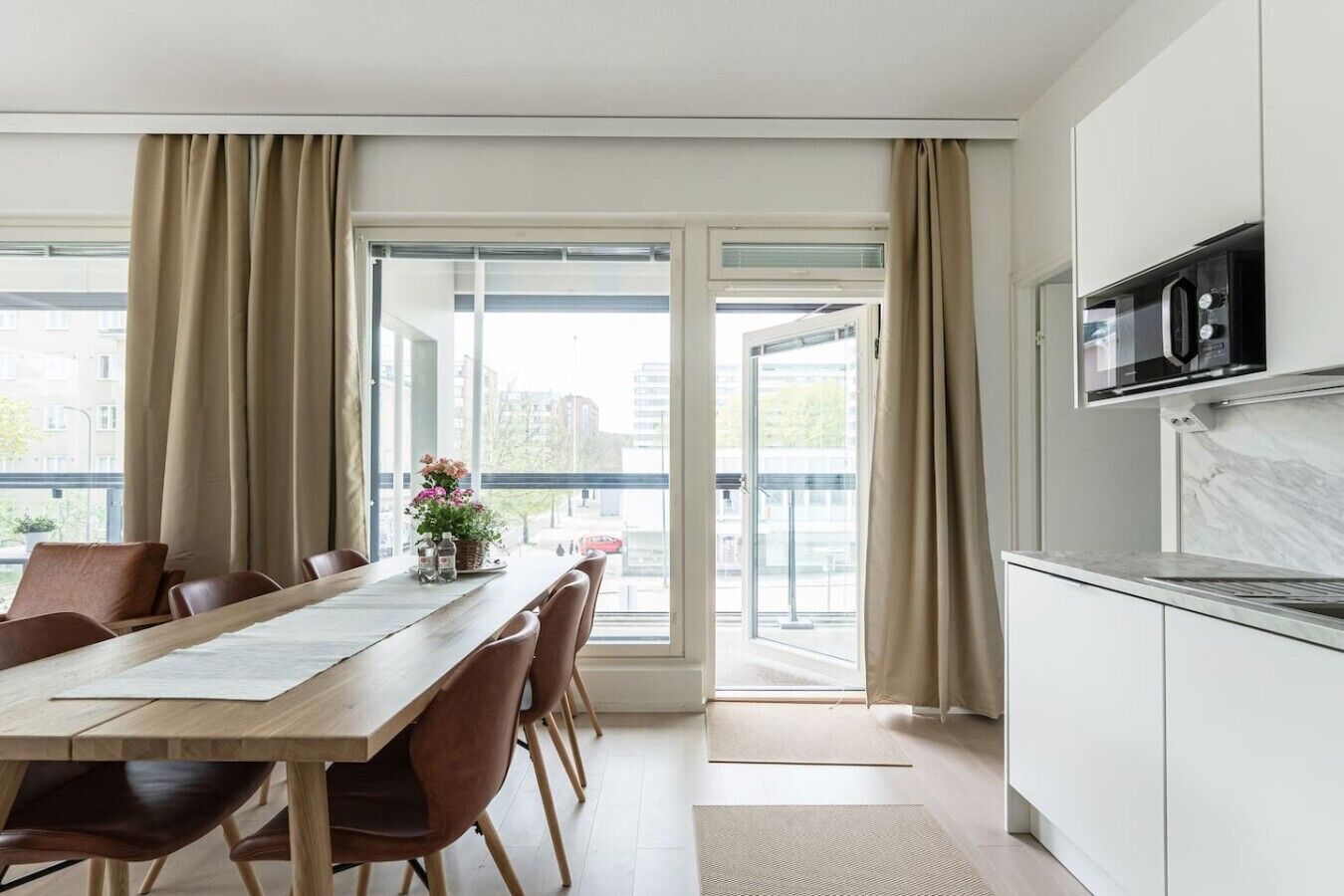 Une cuisine moderne équipée d’un four à micro-ondes, un coin repas avec une table et des chaises en bois, et de grandes fenêtres avec des rideaux.