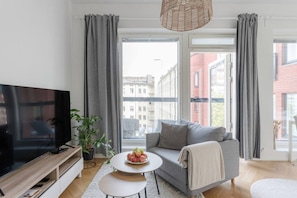 A modern living room with a flat-screen TV, grey sofa, round coffee table with fruit, and large windows with curtains.
