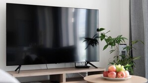 A modern living room with a flat-screen TV on a stand, a small round table with fruit, and a potted plant.