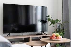 A modern living room with a flat-screen TV on a stand, a small round table with fruit, and a potted plant.